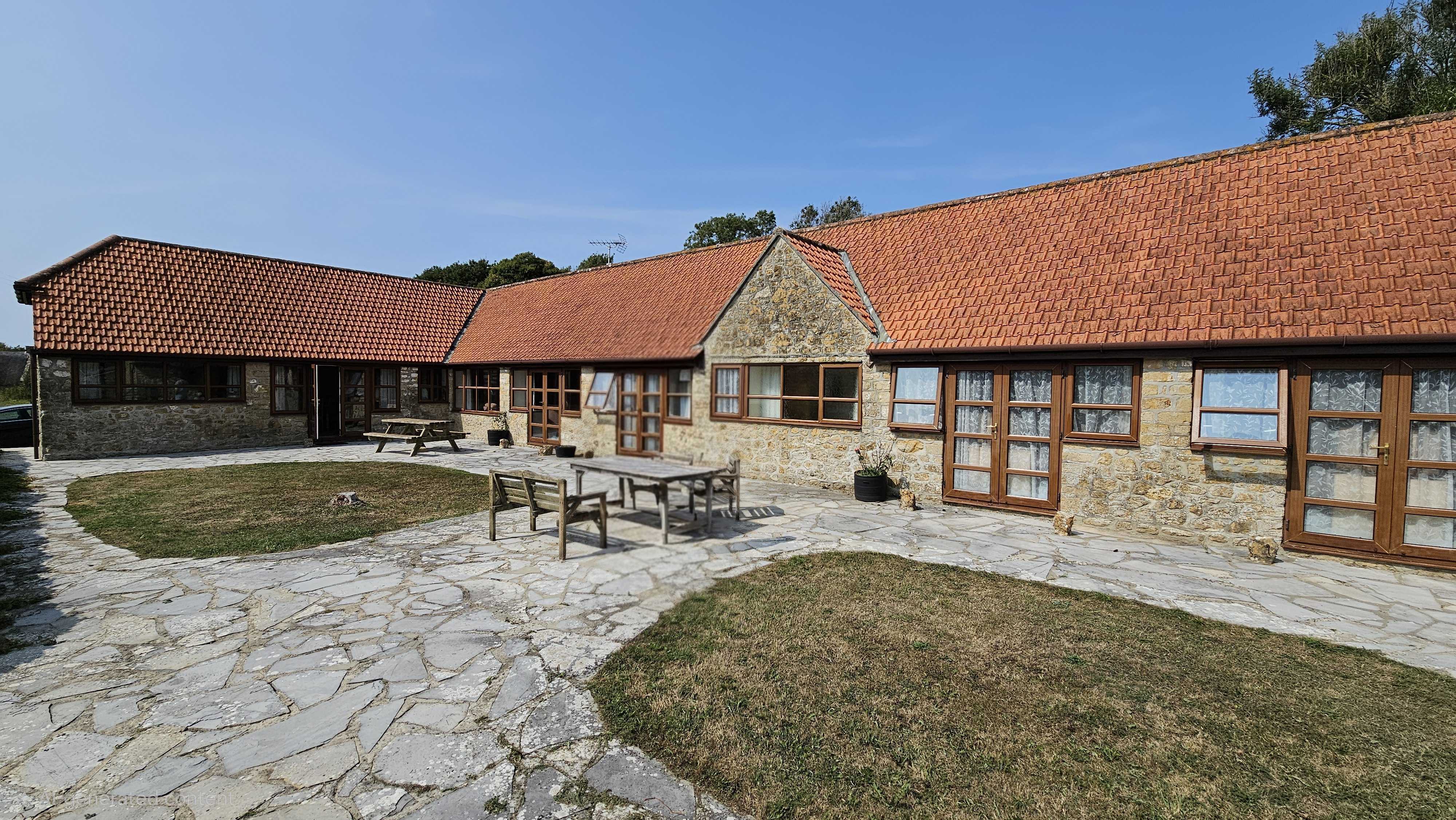 The Old Piggeries - courtyard view showing the long stone-built single-storey cottage with red-tiled roof, picnic bench and flagstone patio.